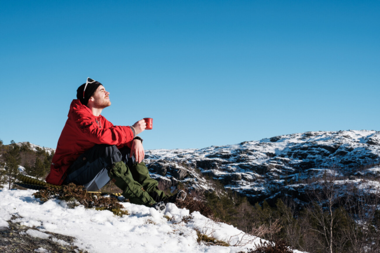 Mann sitter på en snøkledd tue og myser mot sola med turkopp i hånda