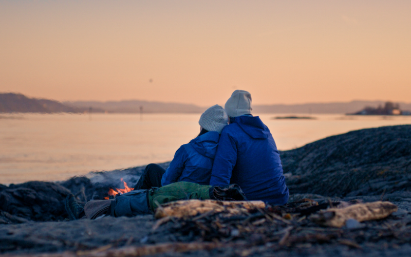 Par sitter rundt et bål ved sjøen.