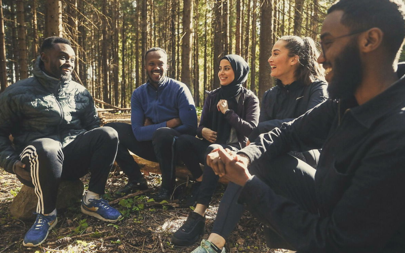 Fem voksne sitter ute i skogen. De ler av en som forteller noe morsomt.