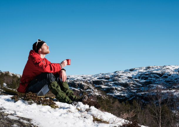 Mann sitter på en snøkledd tue og myser mot sola med turkopp i hånda
