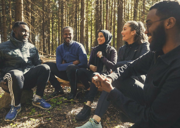 Fem voksne sitter ute i skogen. De ler av en som forteller noe morsomt.